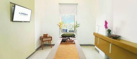 A room with a massage table, a TV mounted on the wall, a window with a view, and a reception desk.