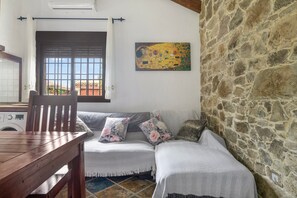 A room with a wooden table, a sofa with floral cushions, a stone wall, and a painting on the wall. - El Cortijo 1 Apartment: Pool, Terrace & AC in Conil de la Frontera (Conil de la Frontera)