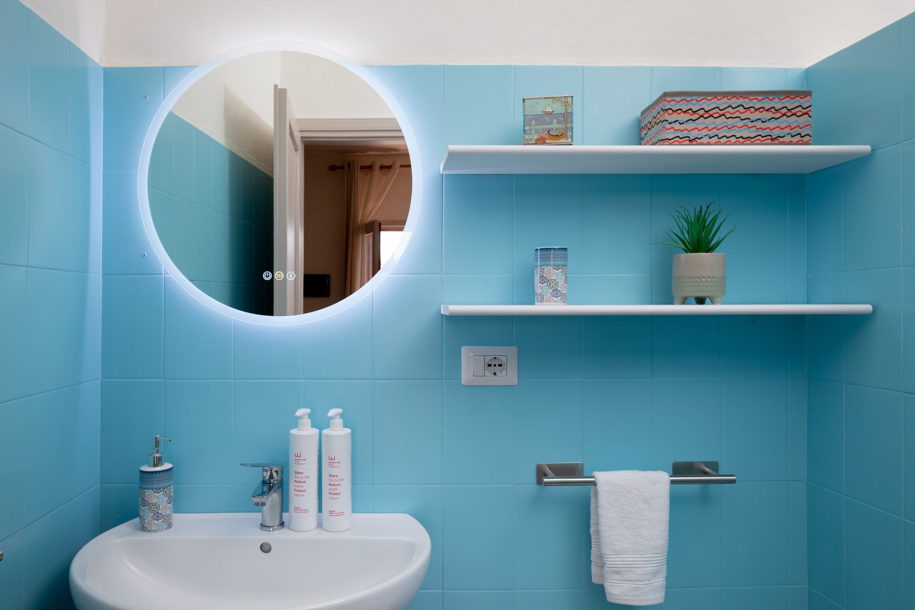 A bathroom with a white sink, a mirror, and blue tiles. There are two shelves with decorative items and a potted plant.