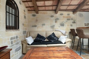 A stone-walled room with a wooden ceiling, a beige sofa with black cushions, a wooden coffee table, and two bar stools.