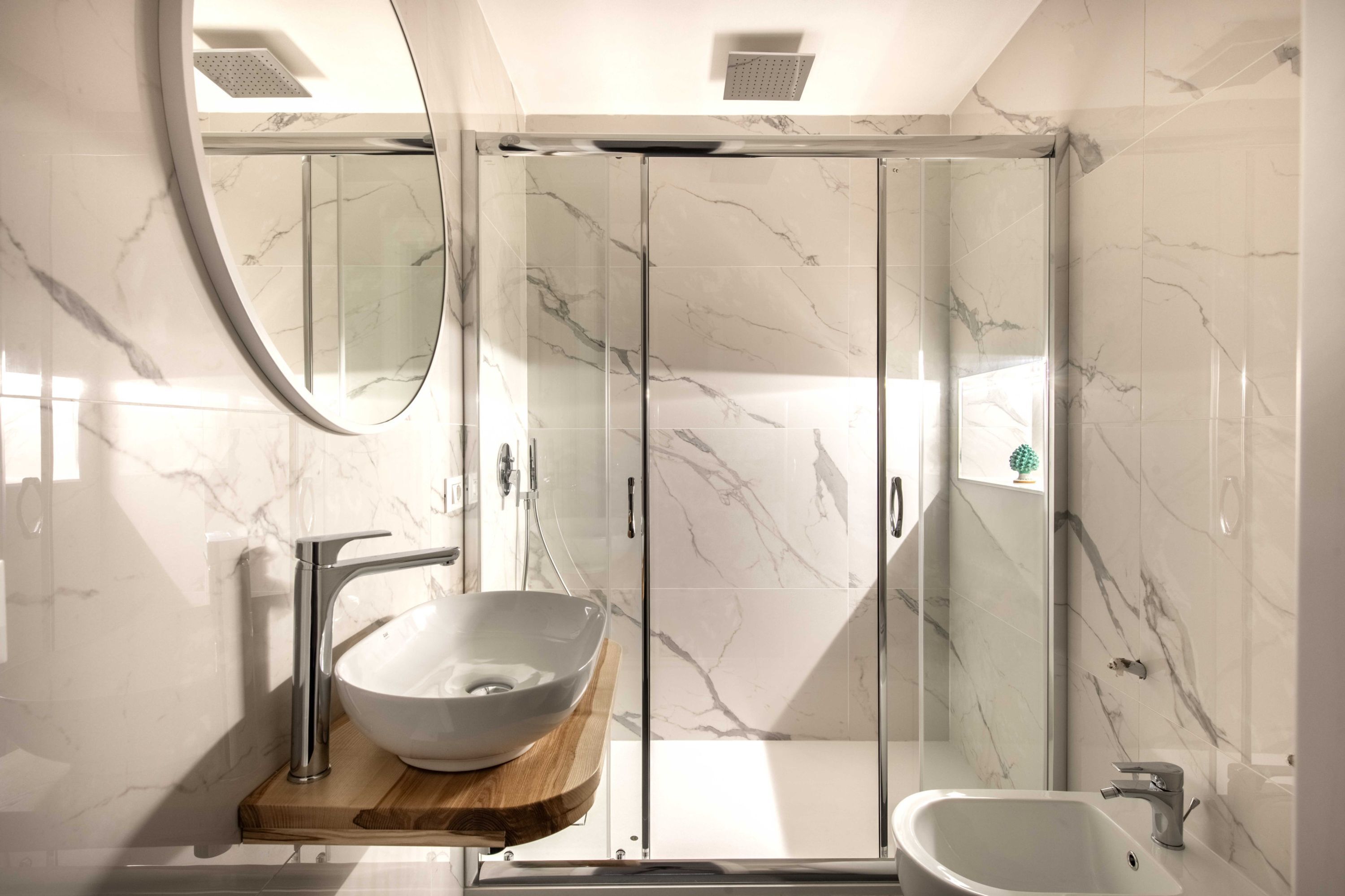 A modern bathroom with a marble backsplash, a glass shower enclosure, a wooden vanity, and a freestanding bathtub.