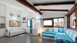 Una sala de estar acogedora con un sofá blanco, un sofá azul y una mesita con una lámpara. Se ve una ventana con cortinas y vistas a árboles.