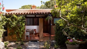 Una casa rústica con techo de tejas, un jardín con diversas plantas y una maceta grande, y un patio con mesa y sillas.
