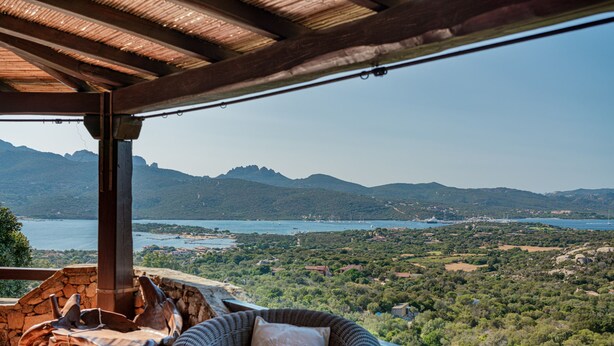 Eine Terrasse mit Korbstuhl und Tisch, mit Blick auf eine reizvolle Landschaft mit Bergen und einem See.
