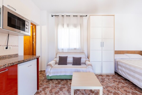 A compact room with a bed, a small table, a kitchenette with a microwave, and a wardrobe.