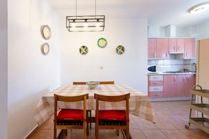 A dining area with a checkered tablecloth, wooden chairs with red cushions, a bowl on the table, and a kitchen in the background with pink cabinets.