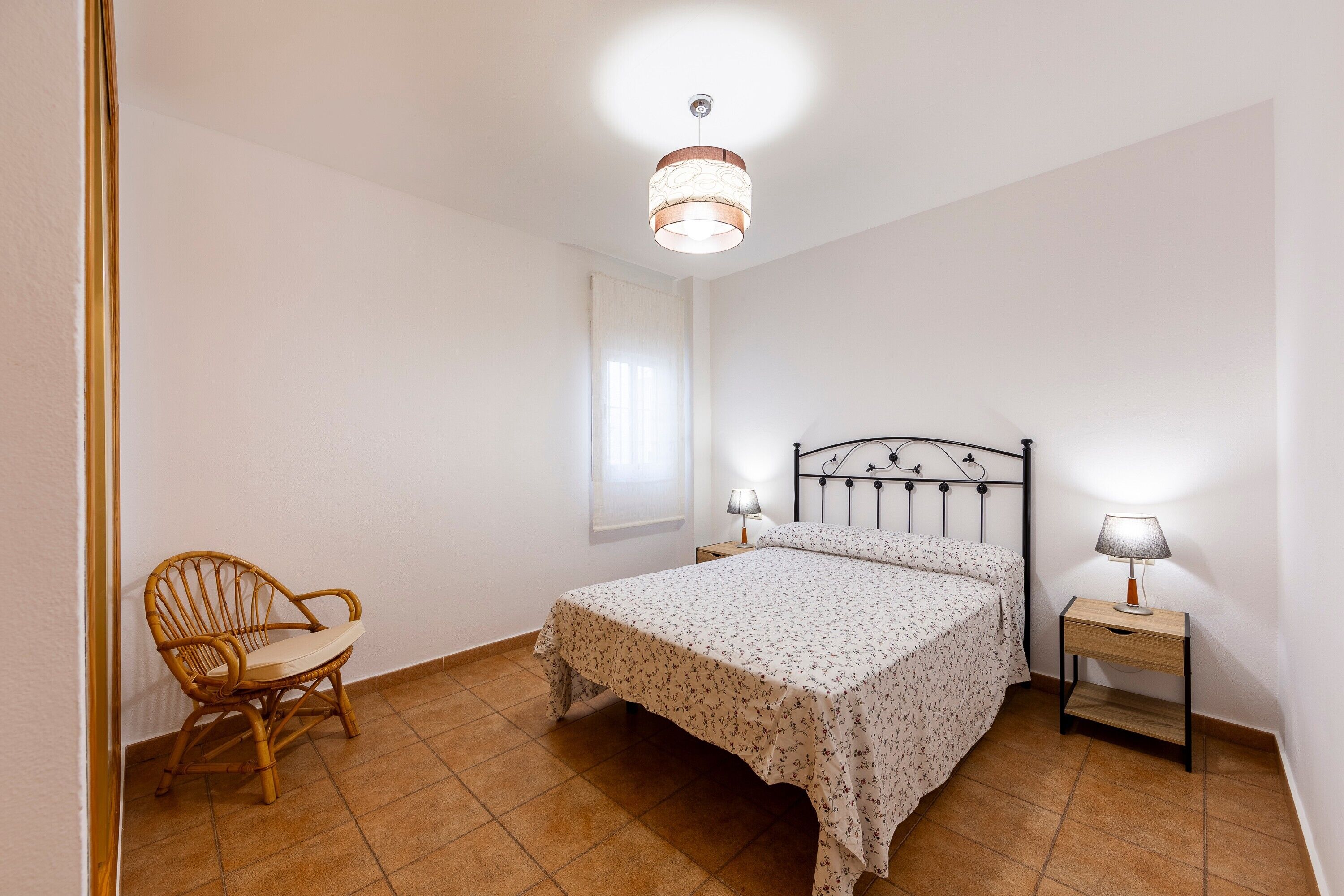 Een slaapkamer met een bed, een stoel, een nachtkastje, een lamp en een plafondlamp.