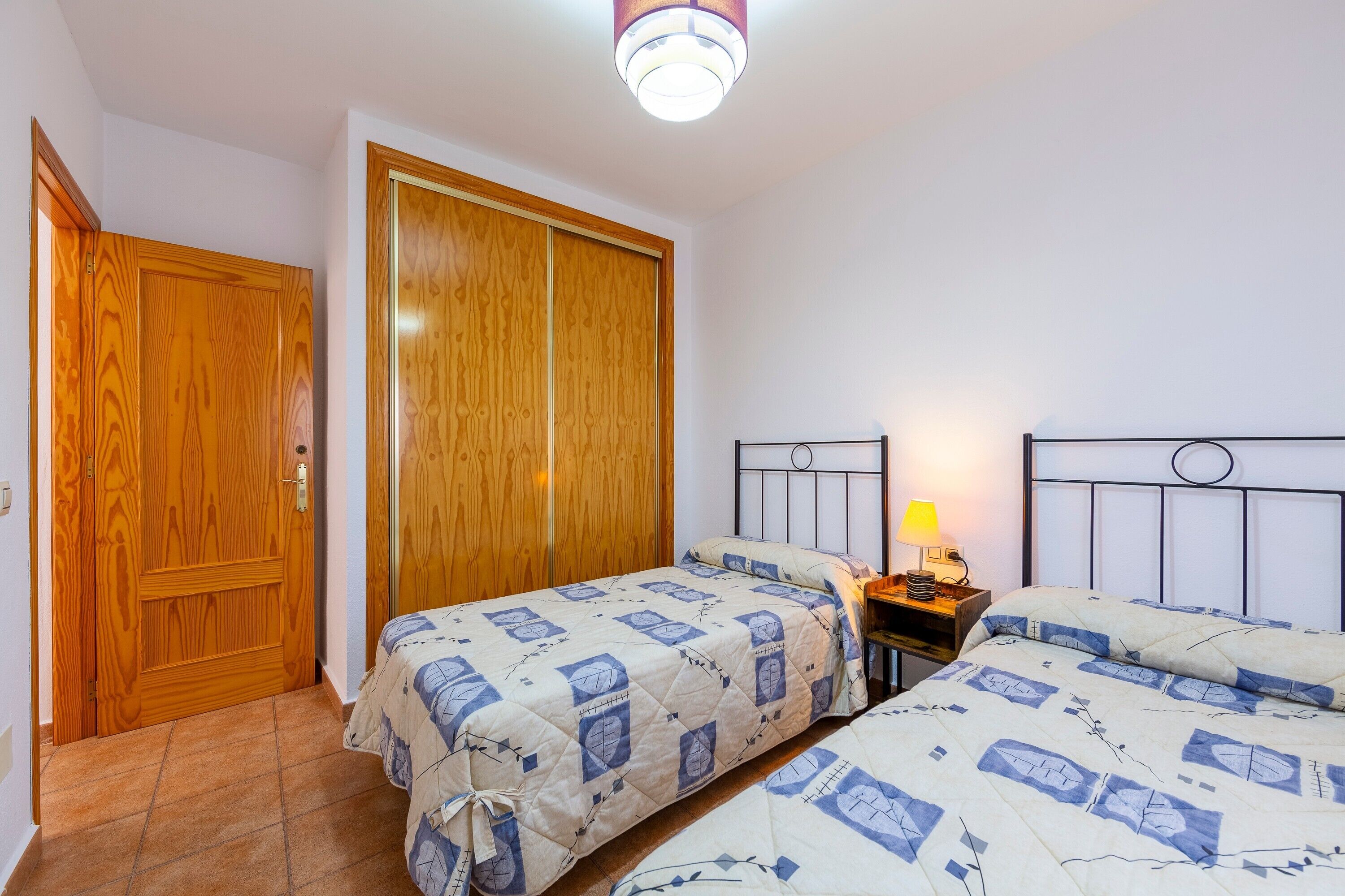 A bedroom with two beds, a wooden wardrobe, a bedside table with a lamp, and a ceiling light fixture.