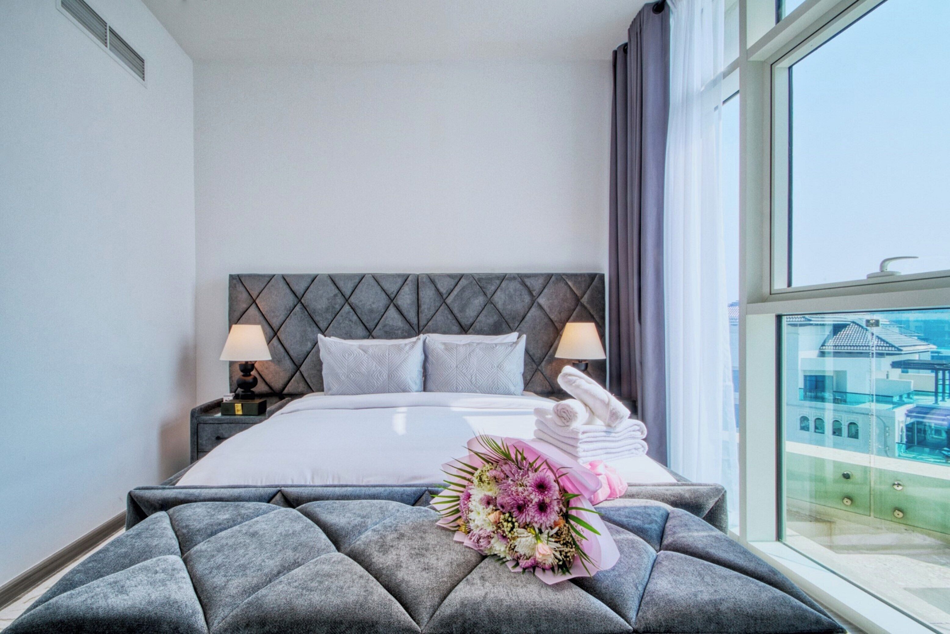 Hotelkamer met een bed, kussens, nachtkastje, lamp, raam met uitzicht en een bloemenboeket op het bed.