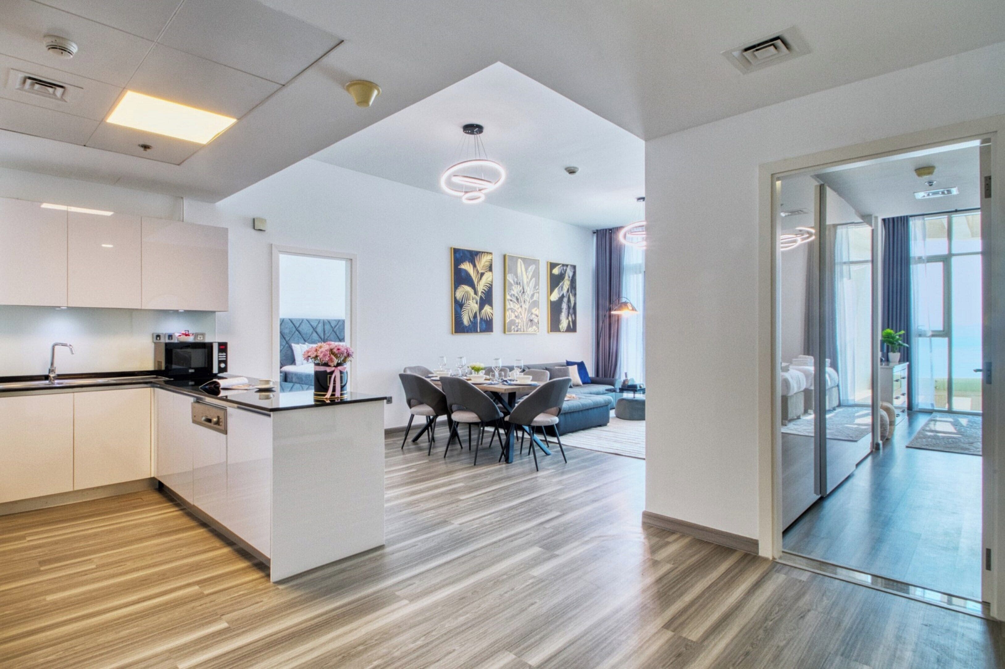 A modern kitchen with a dining area, a living room with a sofa, and a glass door leading to a balcony.