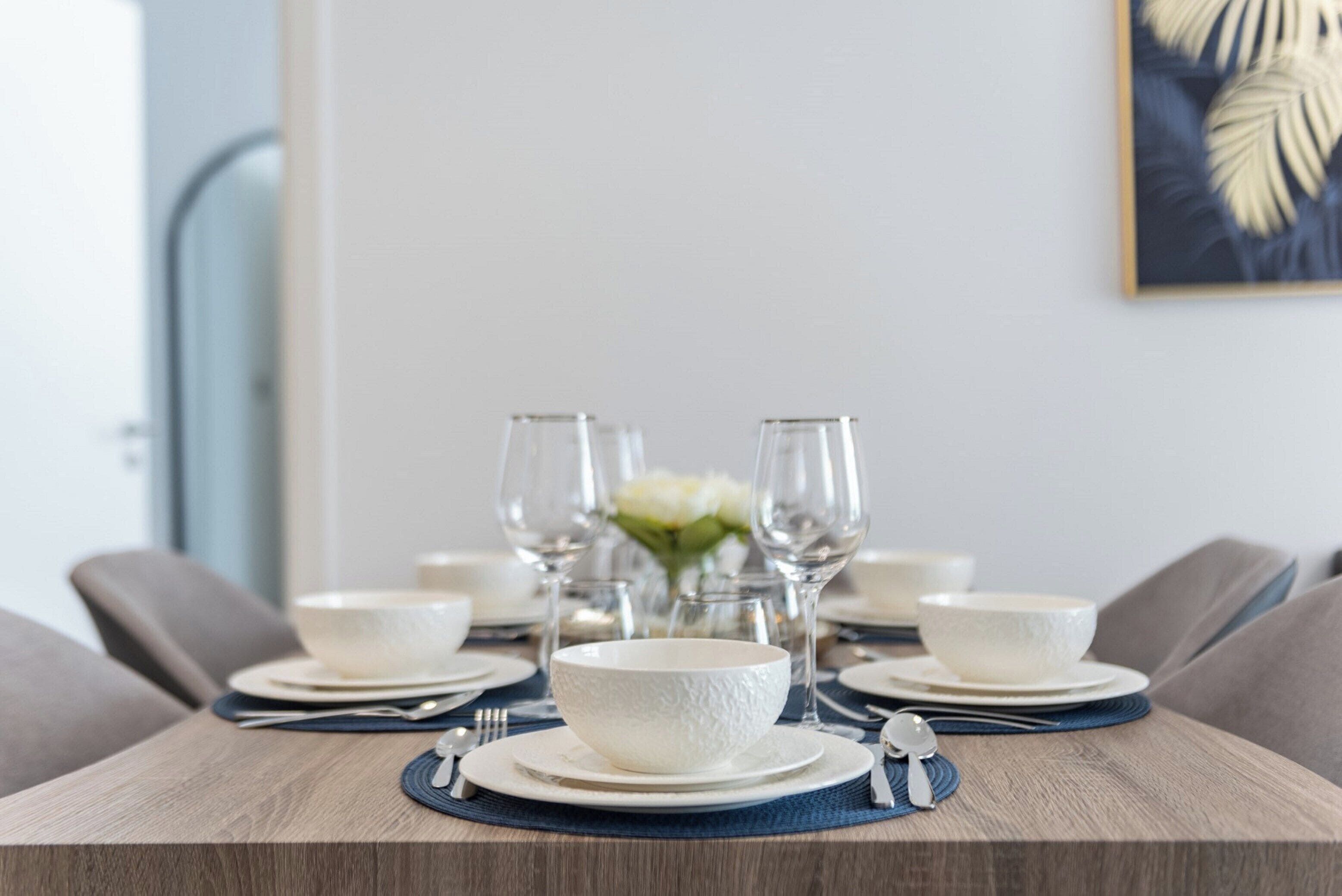 Mesa de jantar posta com tigelas, pratos e talheres brancos, e um vaso de flores ao centro.
