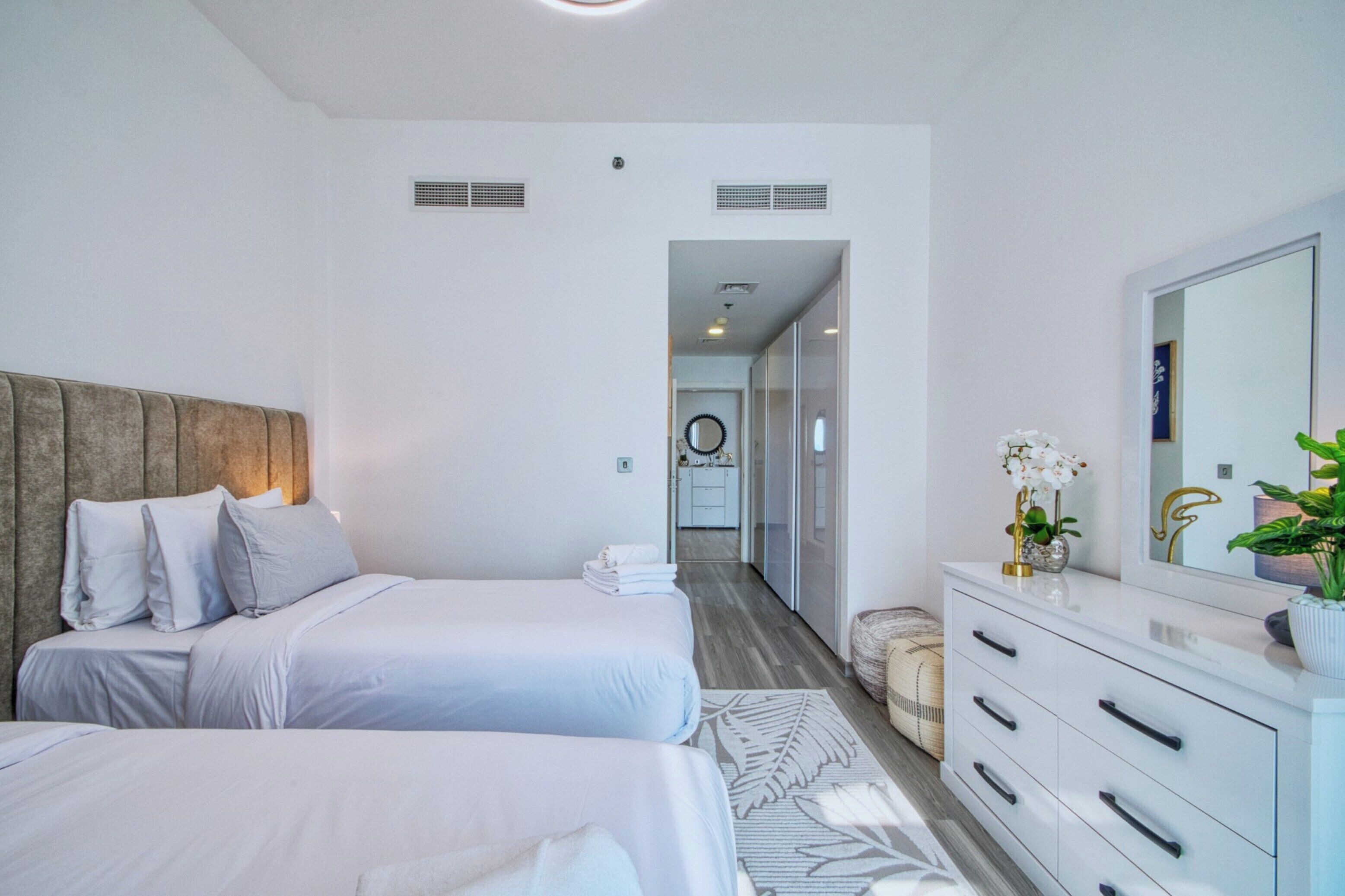 A hotel room with two beds, a large mirror, a vanity with drawers, and a hallway leading to a bathroom.