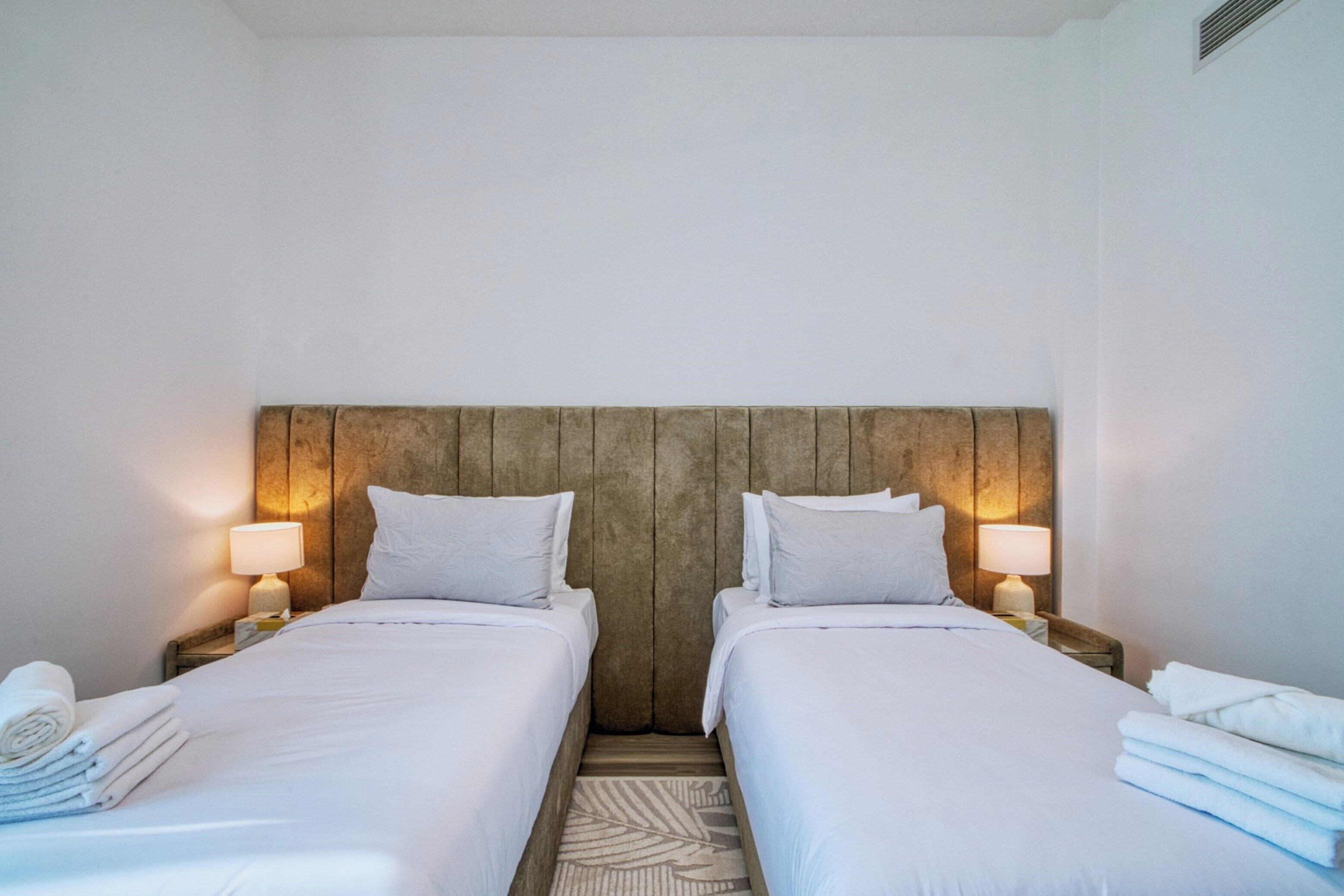 Une chambre double avec deux lits, chacun doté de draps et d’oreillers blancs, d’une tête de lit en bois, de lampes de chevet et d’un tapis sur le sol.