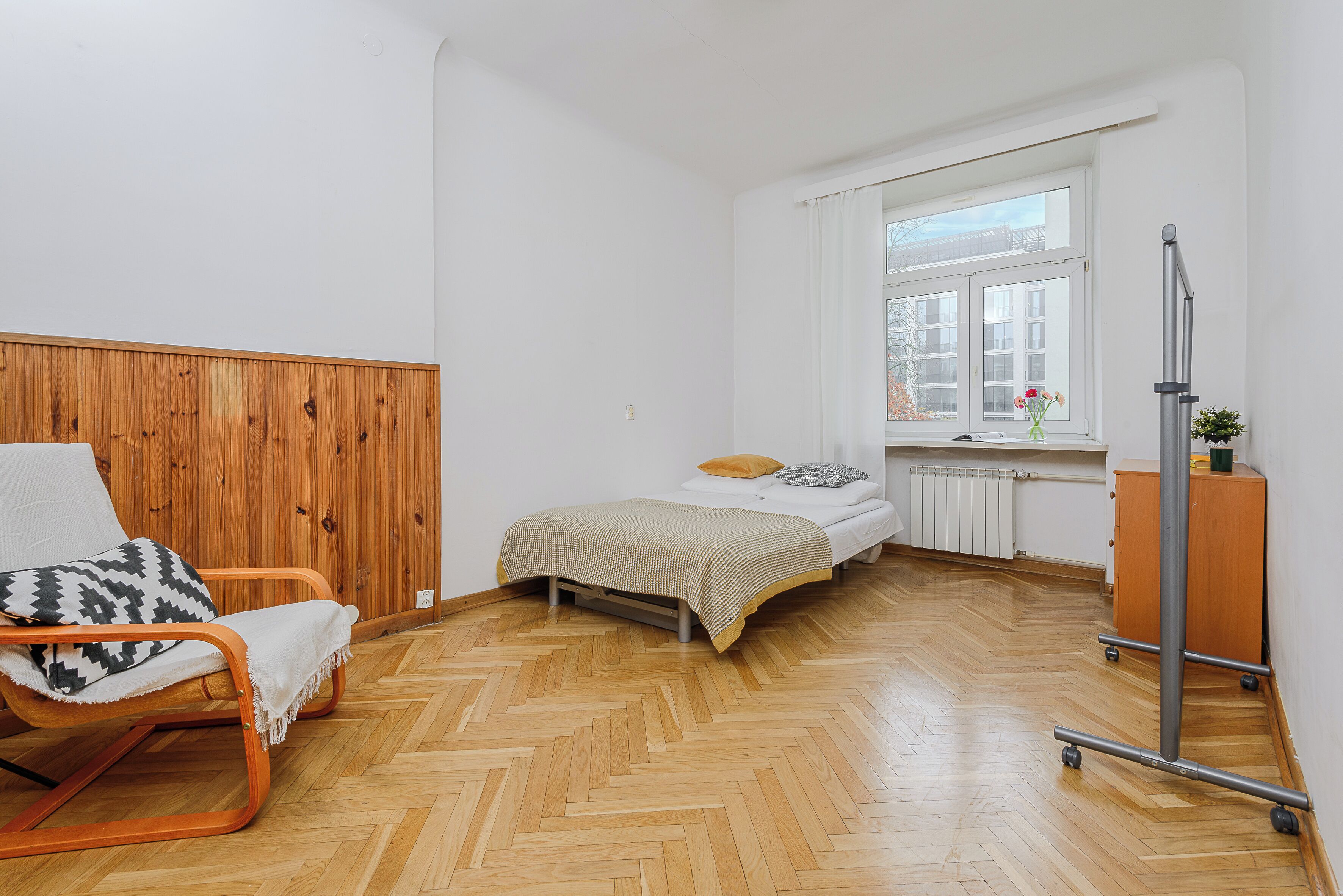 A room with a wooden floor, a bed with a checkered bedspread, a chair with a white blanket, a radiator, and a window with a view of trees.
