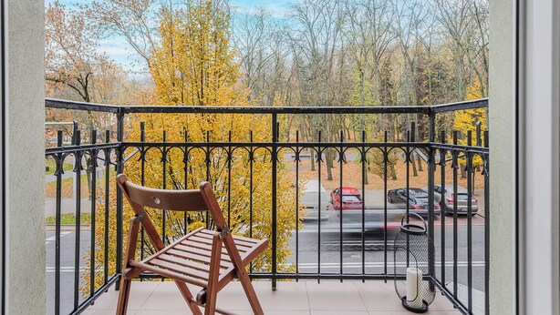 A balcony with a wooden chair, a metal railing, and a view of a street with cars and autumn trees. - ShortStayPoland Szara B170 (Warsaw)