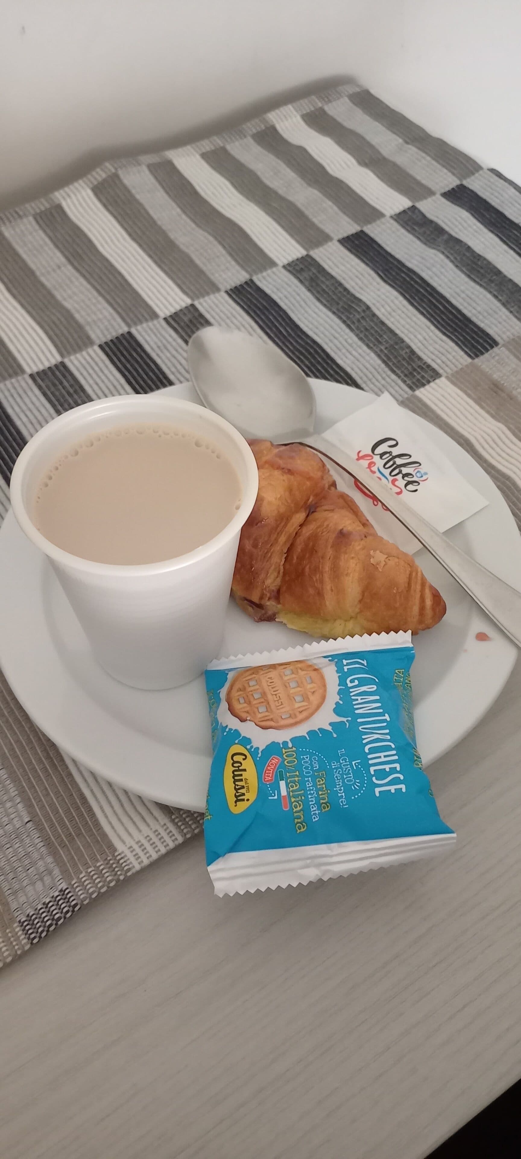 Eine weiße Kaffeetasse, ein Croissant und eine Packung Colosso Kekse auf einem weißen Teller.