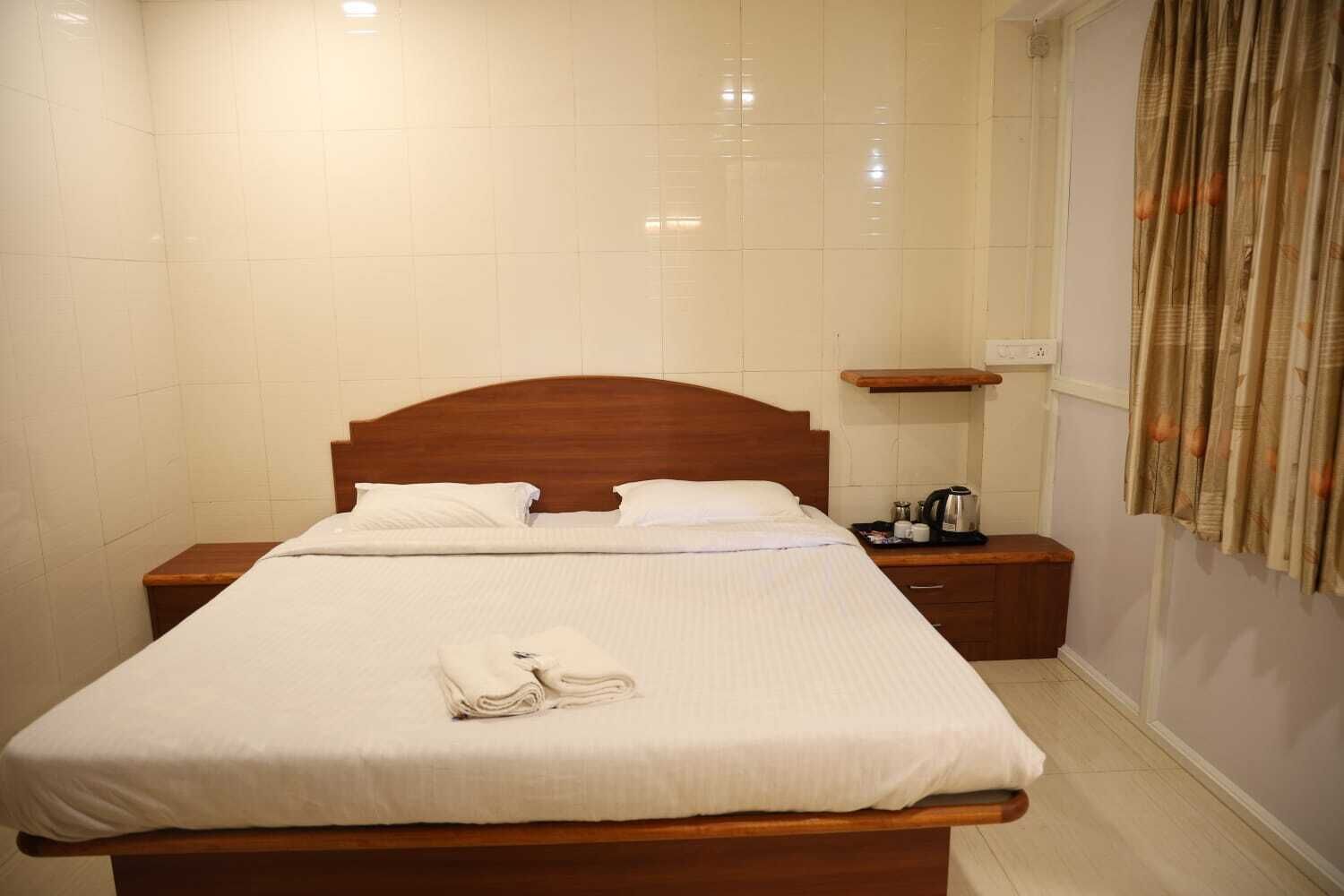 A neatly made bed with white linens and a folded towel, a wooden headboard, a bedside table with a kettle, and a small shelf.
