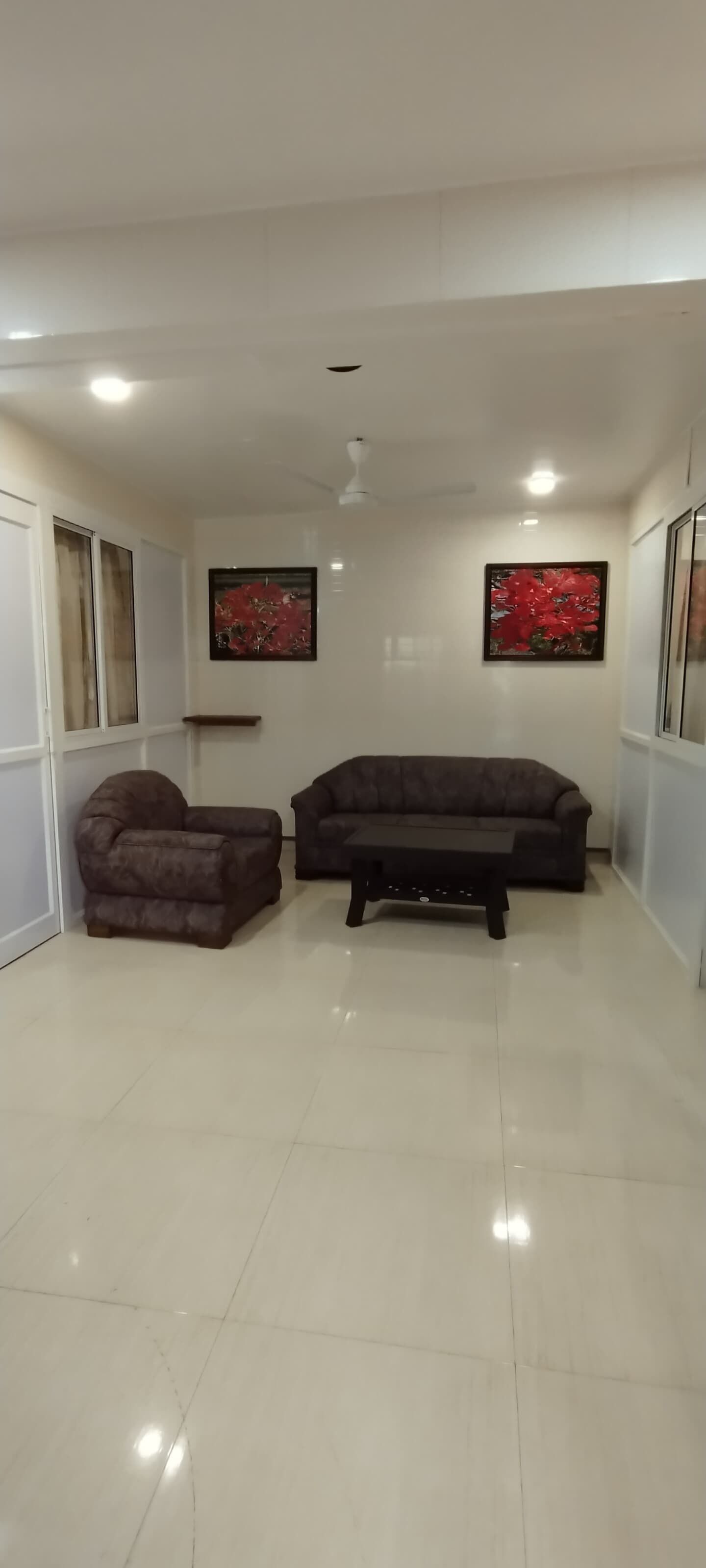 A spacious living room with a dark brown sofa and armchair, a black coffee table, and two red floral paintings on the walls.