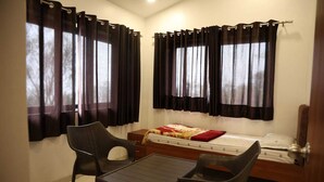 Una habitación pequeña y luminosa con una cama, dos sillas, una mesa y cortinas en las ventanas.