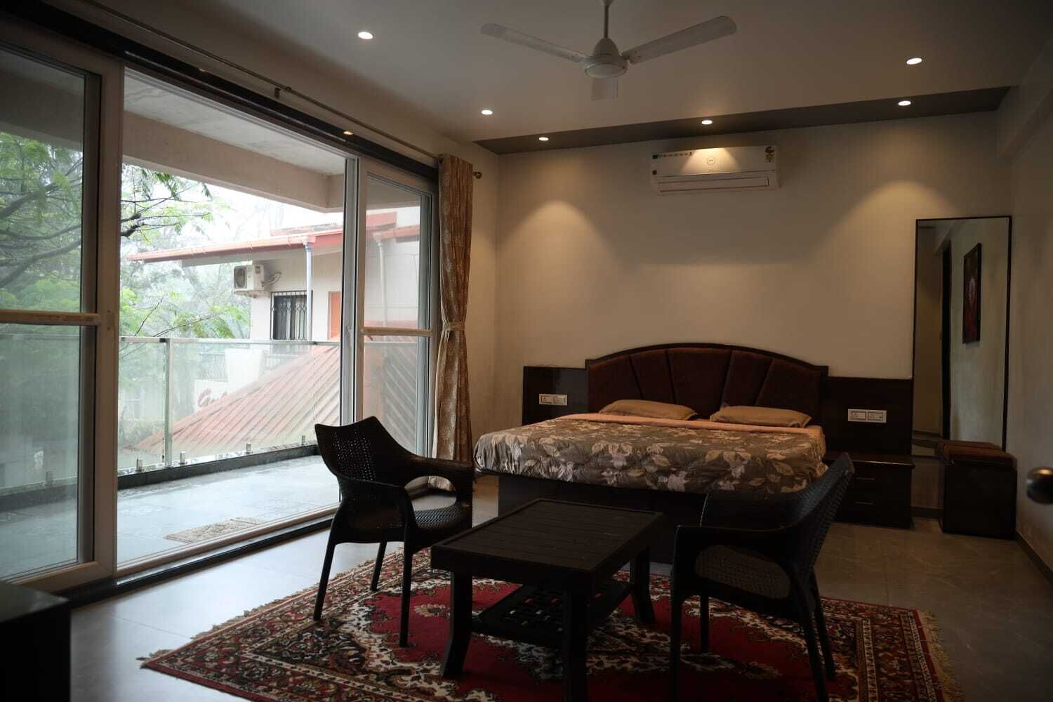 Un dormitorio con cama, escritorio, dos sillas, ventilador de techo y un ventanal con vistas a edificios.
