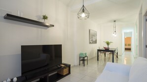 A modern living room with a flat-screen TV, a dining area with a table and chairs, and a hallway leading to a bedroom.