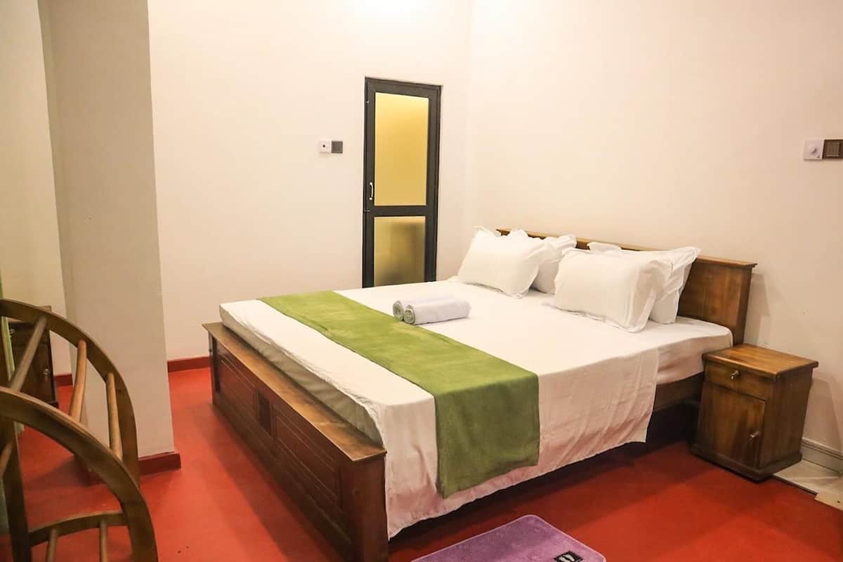 Un lit simple avec une tête de lit en bois, une couverture verte et du linge de lit blanc. Il y a une table de chevet en bois avec un tiroir, un petit miroir et un interrupteur sur le mur.