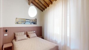 Un dormitorio con una cama, una mesita de noche, una lámpara de pared y un ventanal con cortinas traslúcidas.