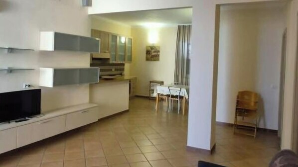 A modern living room with a flat-screen TV, a dining area with a table and chairs, and a kitchen with cabinets and a countertop.