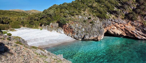 A rocky coastline with clear turquoise waters, a white sandy beach, and dense green vegetation on the hills.