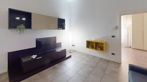 A modern living room with a flat-screen TV, a dark entertainment unit, a yellow shelf with decorative items, and a large window providing natural light. - Charlotte apartment wheels (Firenze)
