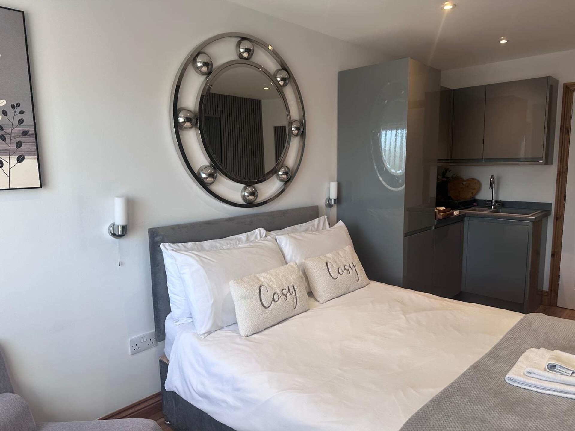 Quarto de hotel com uma cama, um espelho redondo com iluminação, uma TV e uma kitchenette.