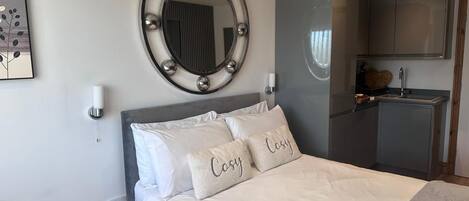 Quarto de hotel com uma cama, um espelho redondo com iluminação, uma TV e uma kitchenette.