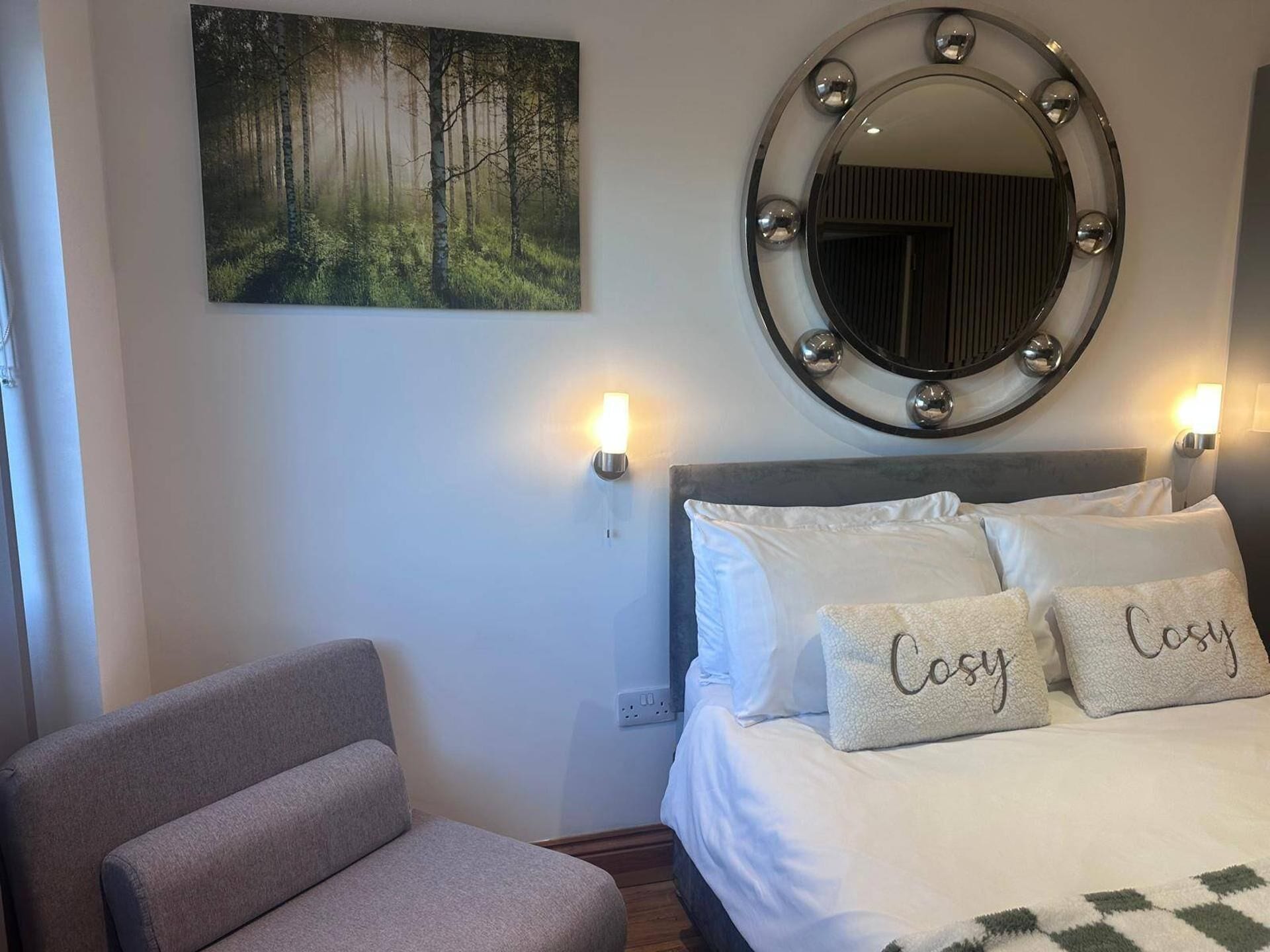 A bedroom with a bed, two bedside lamps, a round mirror, a gray armchair, and a forest-themed wall art.