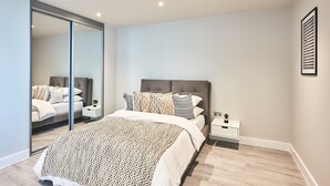 A modern bedroom with a large bed, a nightstand, a mirror, and a wall-mounted light fixture. - Cozy Studio in Bracknell With Free Parking (Bracknell)