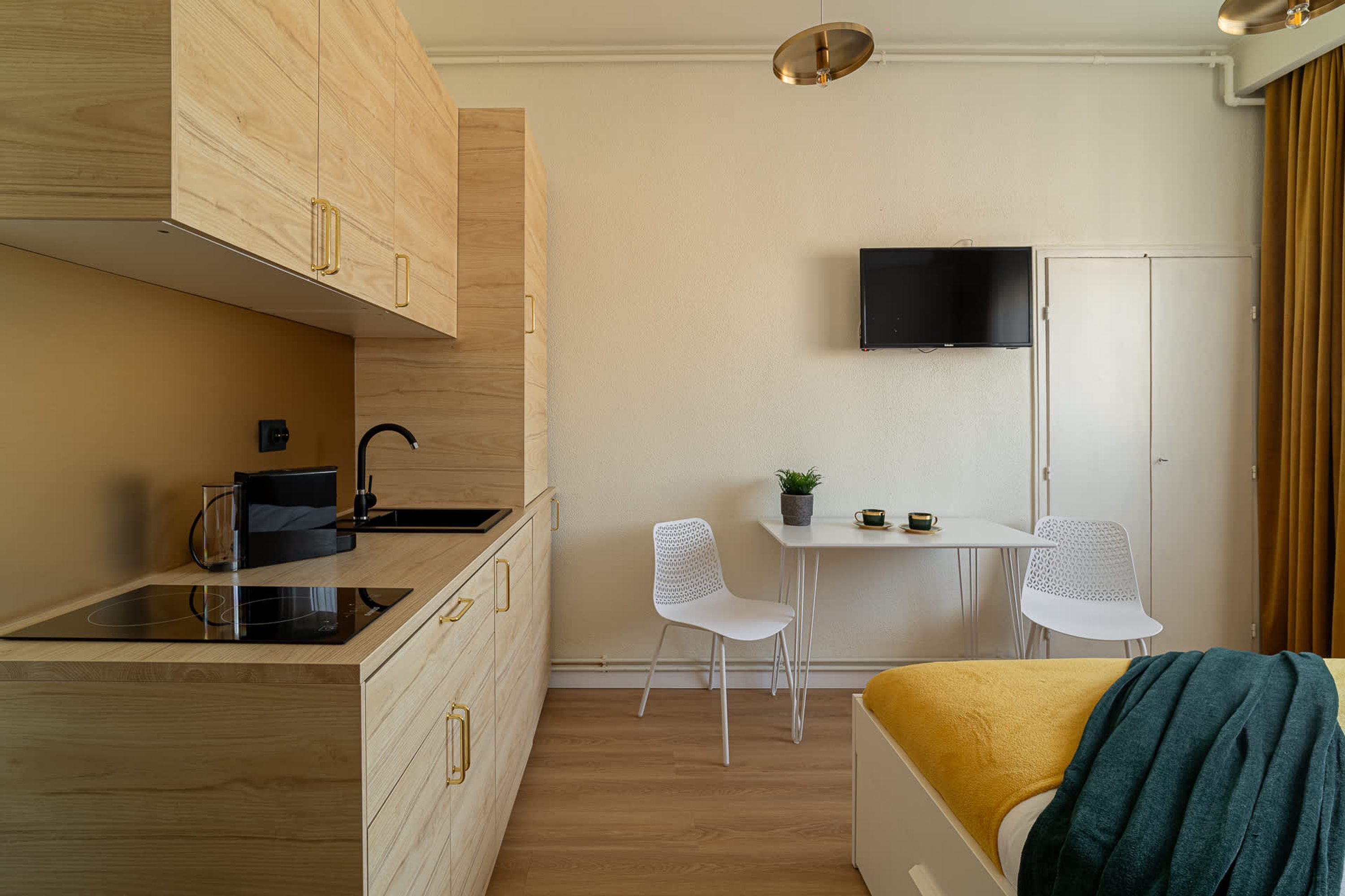 A compact room with a kitchenette, a bed with a yellow blanket, a TV mounted on the wall, and a small table with chairs.