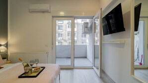 Habitación de hotel con cama, un atril con aperitivos y una botella, televisor montado en la pared y puerta corrediza de vidrio que da a un balcón exterior.