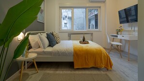 Un dormitorio con una cama, un escritorio, una silla, una planta grande y un televisor.