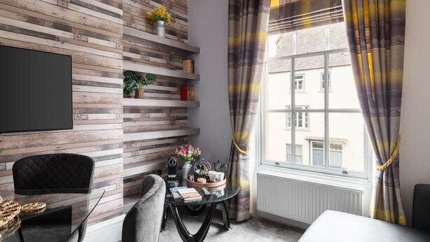 A modern living room with a wooden wall panel, a glass coffee table, a black sofa, and a view of a building through a window with striped curtains. - Host Stay Mayford Apartment (Durham)