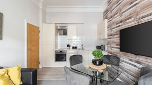 A modern living room with a glass dining table, grey upholstered chairs, a yellow throw pillow, and a flat-screen TV mounted on a wooden wall. - Host Stay Mayford Apartment (Durham)