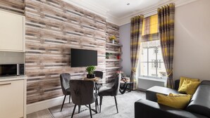 A modern living room with a wooden wall, a flat-screen TV, a dining area with chairs, a sofa with cushions, and a window with curtains. - Host Stay Mayford Apartment (Durham)