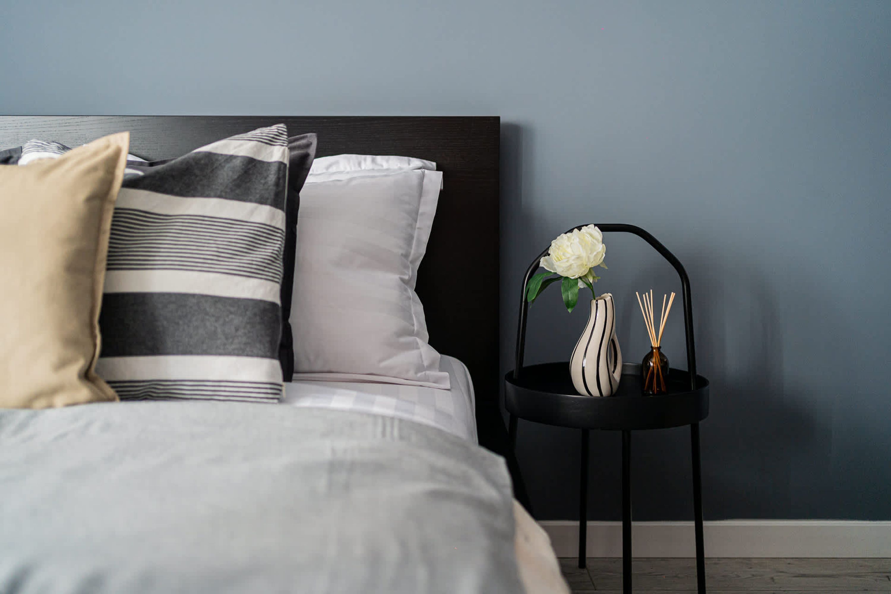 Een netjes opgemaakt bed met gestreepte en effen kussens, een klein zwart nachtkastje met een bloemenvaas, en een donkerblauwe muur.