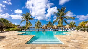 A swimming pool with palm trees and thatched umbrellas, surrounded by lounge chairs and a clear blue sky. - Stylish Apartment With Pool Near Jan Thiel Beach (Jan Thiel)