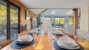 Un espace repas avec une longue table dressée pour le repas, de grandes fenêtres et une vue sur l’extérieur.