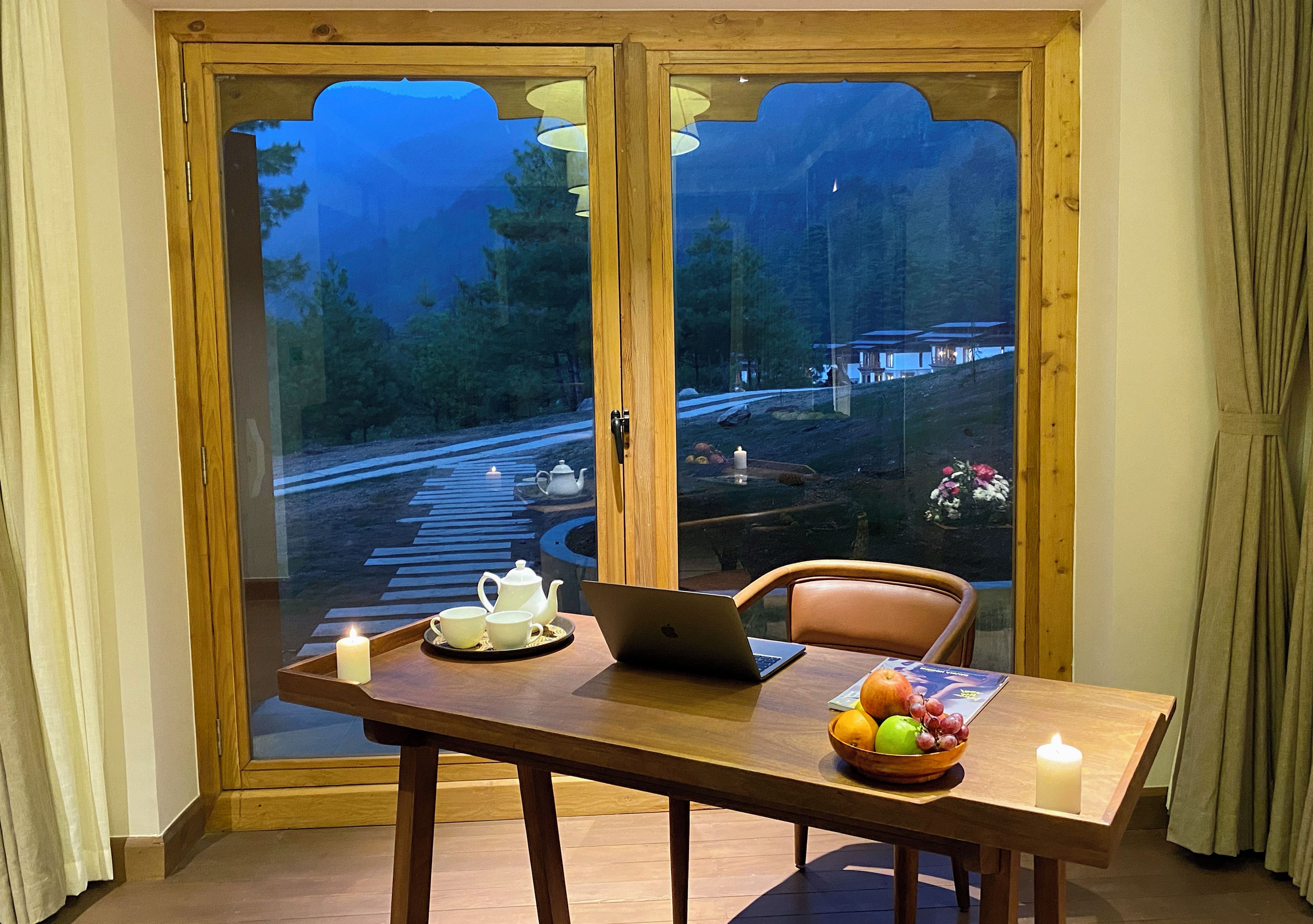 A dining table with a laptop, a candle, a tray with a teapot and cups, and a bowl of fruit.