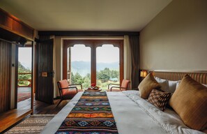 A hotel room with a bed, a chair, a table, and a view of mountains through a large window. - Ozen Resort (Paro)