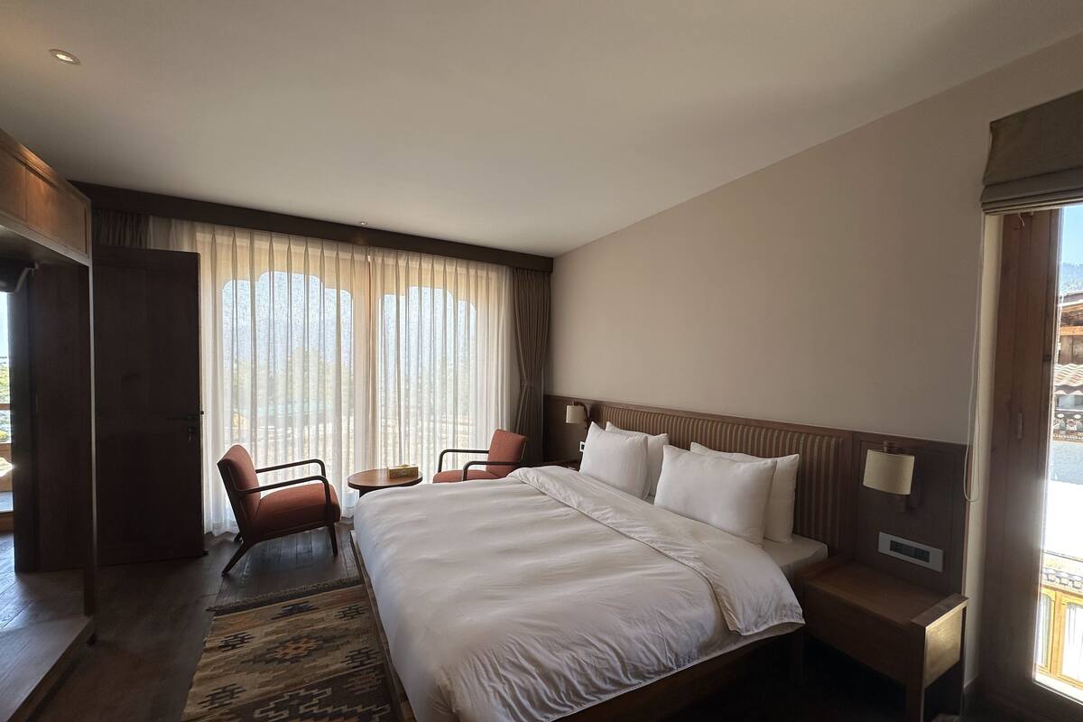 Une chambre d’hôtel avec un grand lit, deux tables de chevet, un bureau, une chaise et une vue sur l’extérieur à travers des rideaux légers.