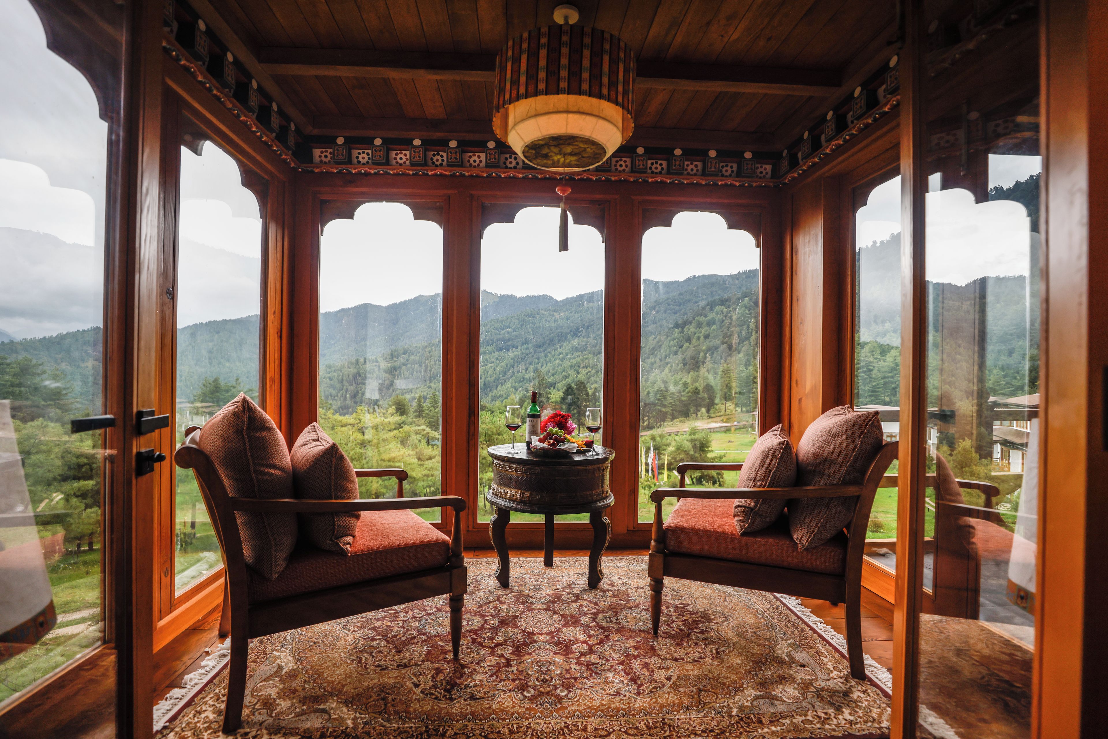 A room with a wooden ceiling, a chandelier, two chairs with cushions, a table with a bottle and glass, and a view of a mountainous landscape through large windows.