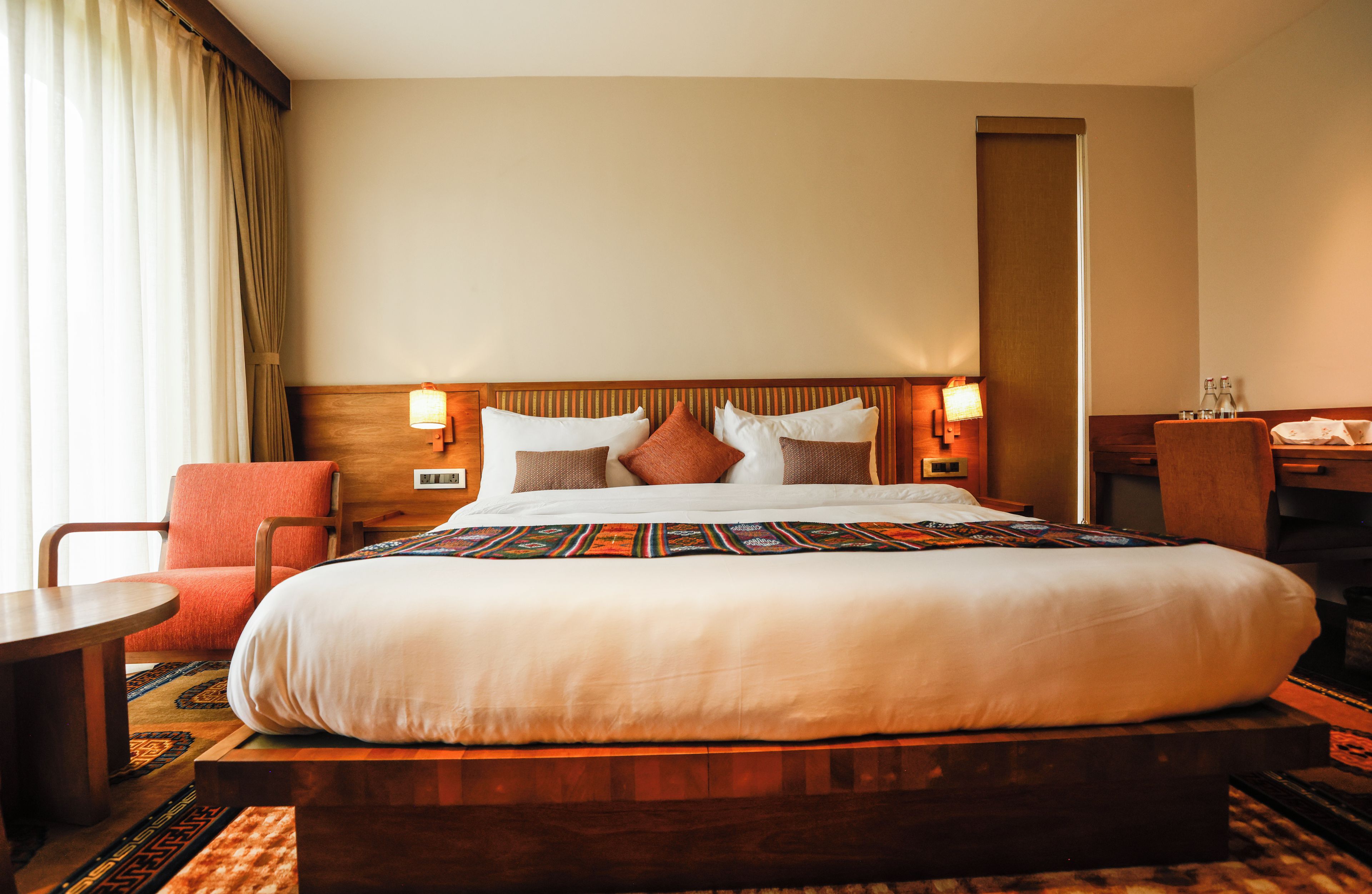 Habitación de hotel con una cama grande, cabecera de madera, dos lámparas de noche, un sillón rojo, una mesita pequeña y piso alfombrado.