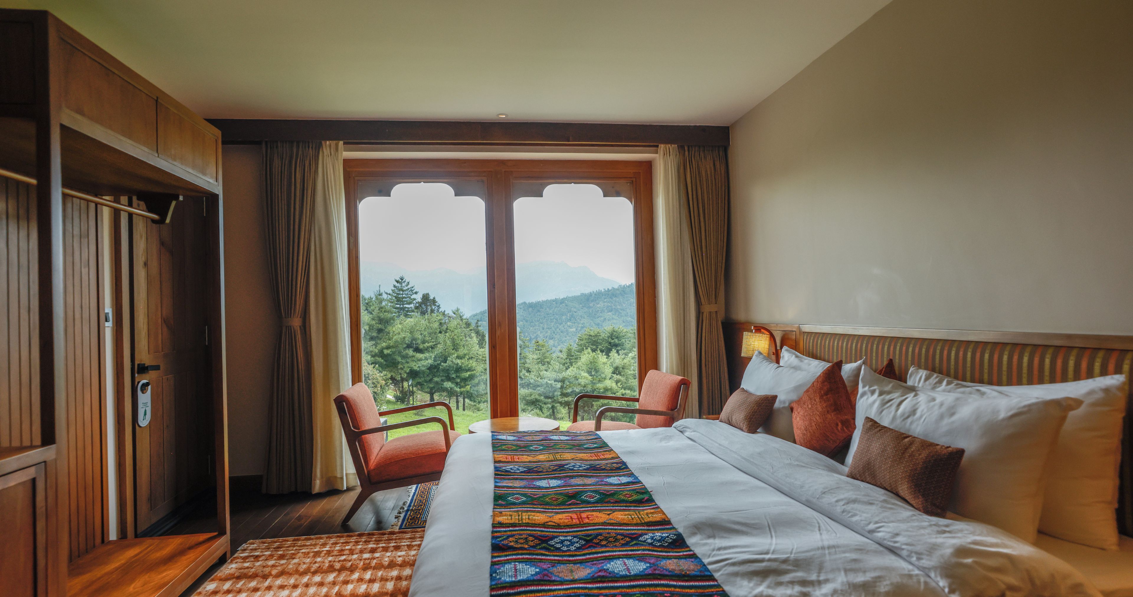 Habitación de hotel con cama, dos sillas, un armario de madera y un gran ventanal con vista a la montaña.