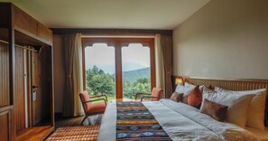 Habitación de hotel con cama, dos sillas, un armario de madera y un gran ventanal con vista a la montaña.